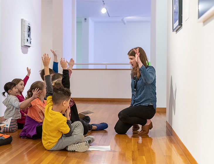Children sitting on the floor in an art gallery raising their hands in response to a question by the docent speaking in front of them.