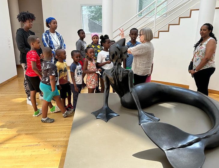 Group of young school kids on a tour at the art museum. A docent is talking to the kids about a sculpture in front of them, Wangechi Mutu's "Water Woman"