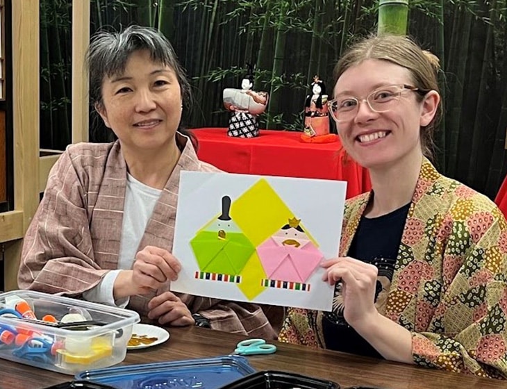 Two people sitting at a table, smiling at the camera and displaying their Japanese origami project.