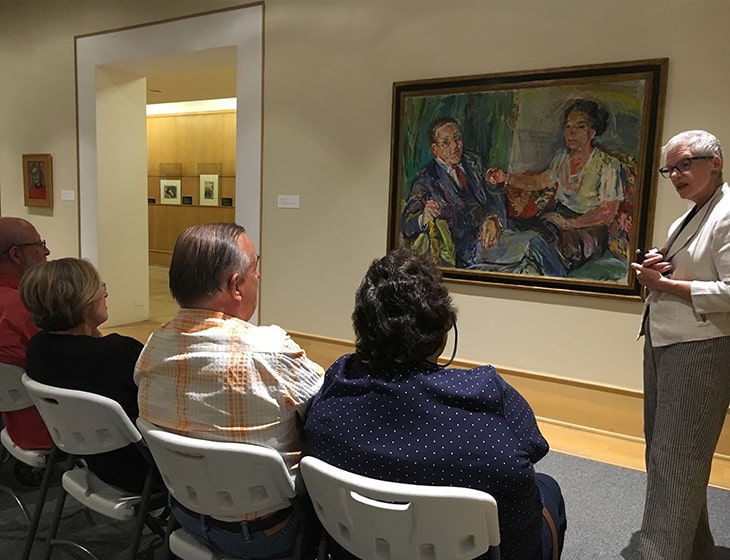 Group of adults gathered in folding chairs in an art gallery. A docent is leading a discussion about a painting.