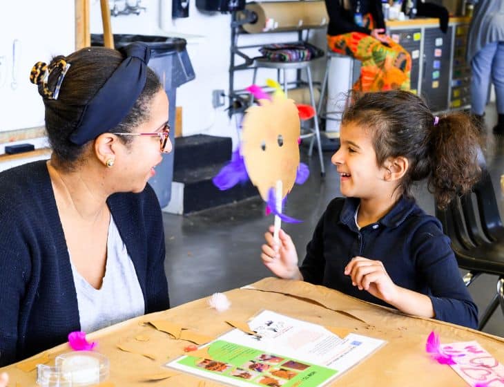 Adult and child sitting at a table in an art studio. Child looks at adult holding up their artwork and smiling.