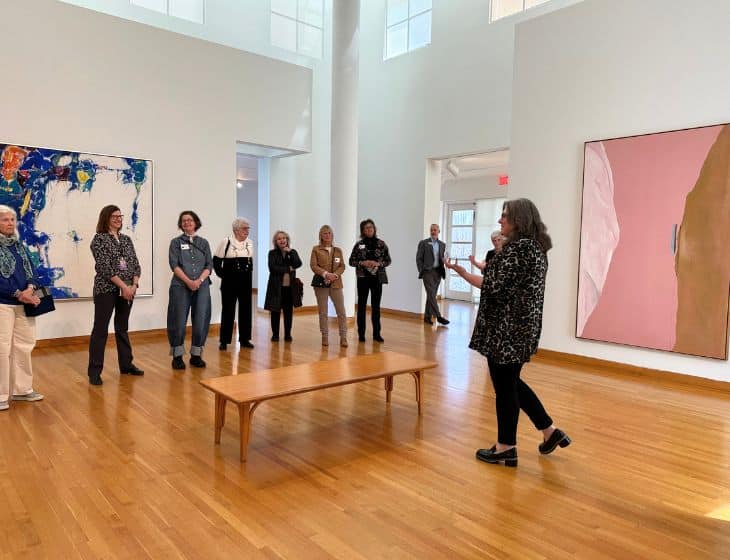 Group of people in a brightly lit gallery with two large artworks on the wall. A curator leads a discussion about the artworks.