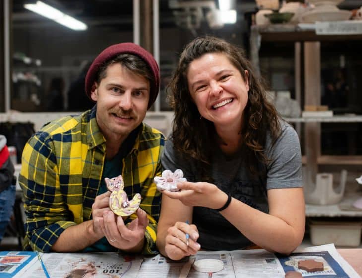 Two people sitting at a table making holiday crafts. They are both looking up at the camera and smiling.