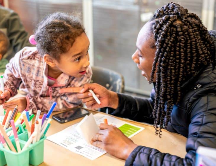 A child and adult sitting at a table working on an art project. They are looking at each other with an expression of surprise.