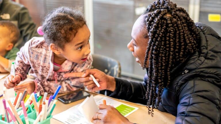 A child and adult sitting at a table working on an art project. They are looking at each other with an expression of surprise.