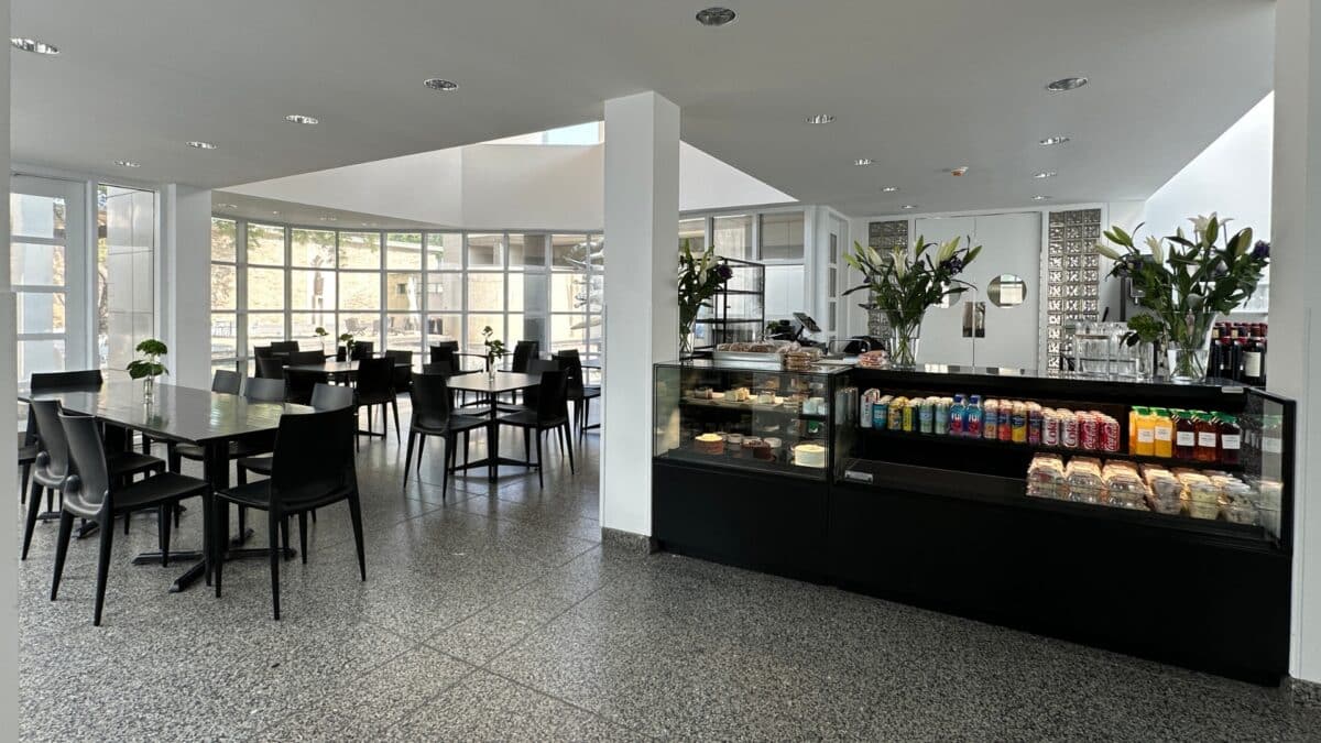 The Art Center cafe presented by la mie. A room with lots of windows overlooking the Art Center courtyard. Black tables and chairs are set up next to the food counter and display case.