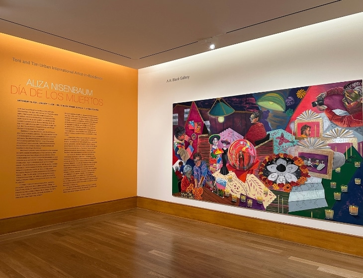 Gallery image of “Aliza Nisenbaum: Día de los Muertos” featuring a large colorful artwork and exhibition text vinyl on a golden painted wall.