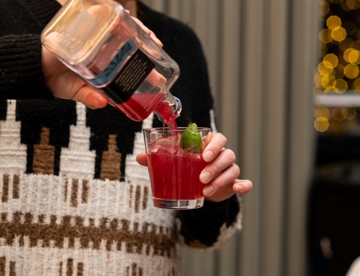 Person in a holiday, sparkling sweater pouring a red drink into a glass with a lime garnish.