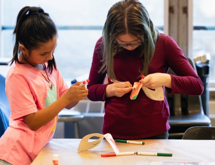 Two young girls in the Art Center studio drawing on eye-face masks