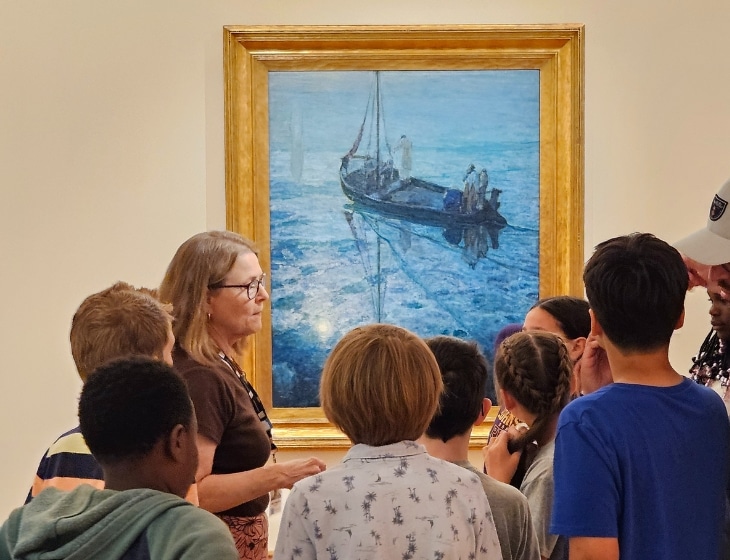 Group of students listening to an art center docent describe an artwork. The art is a blue, sea scape painting with a boat titled, "The Disciples See Christ Walking on the Water" by Henry Ossawa Tanner