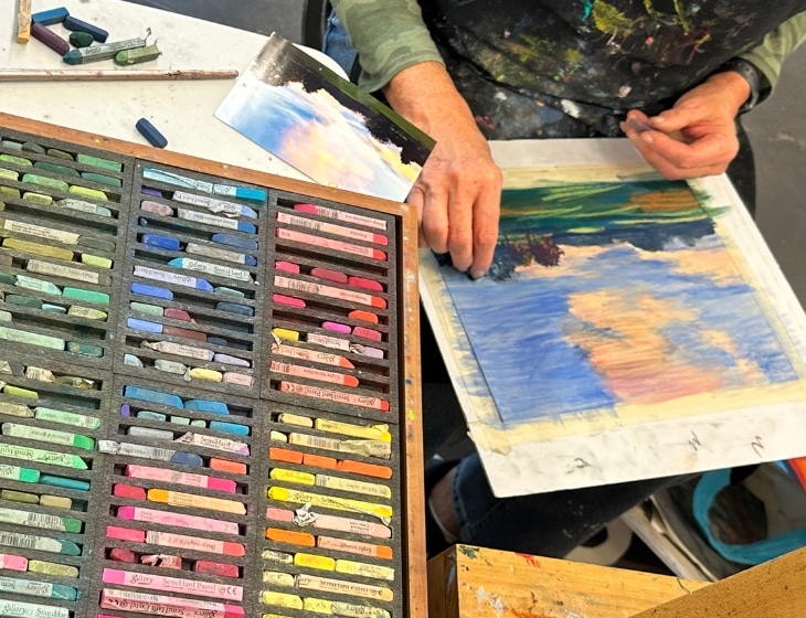 A person's hands working on an artwork. Their canvas sit on their lap and a large pallet of oil pastels sit on the table.