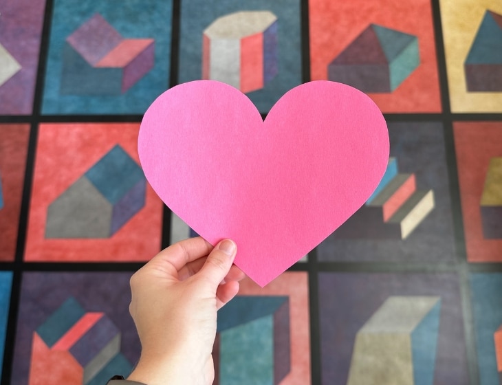 A cut out pink heart held up in front of a large artwork - Sol LeWitt's "Wall Drawing #601, Forms Derived from a Cube (25 Variations)"
