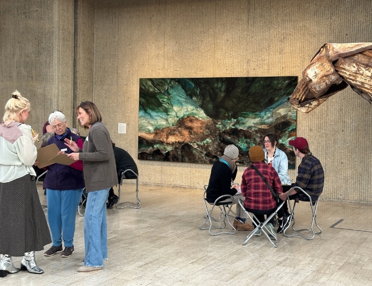 Several people in the Des Moines Art Center galleries, they're talking with each other in small groups. A larger blue/green mosaic artwork hangs on the wall behind them.