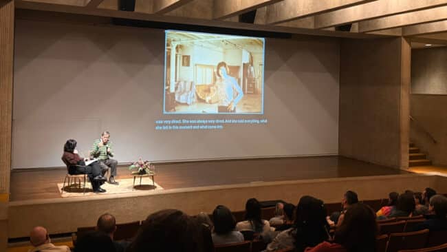 Lecture, "Exploring Maria Lassnig's New York Years," featuring Hans Werner Poschauko and Ashton Cooper - both individuals are sitting on stage talking to each other while Lassnig's artworks and photographs are projected on a large screen.