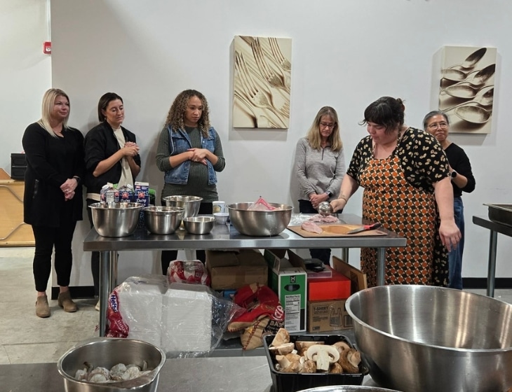 Several people standing and watching a chef prepare ingredients for a meal during a cooking class in a commercial kitchen.